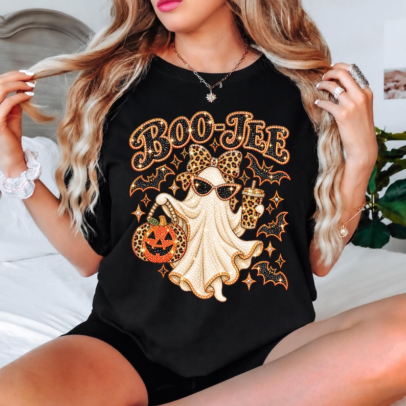 May include: A black t-shirt featuring a graphic of a ghost wearing a leopard print bow and sunglasses. The ghost holds a leopard print pumpkin bucket and a matching cup. The text BOO-JEE is printed above, surrounded by small orange bats and stars.