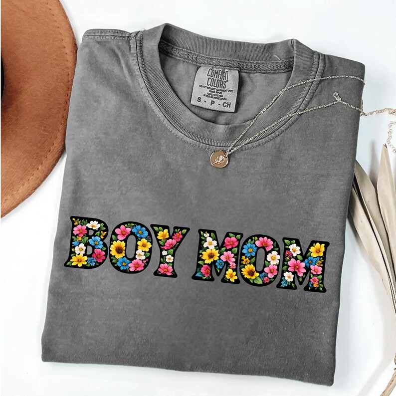 May include: A gray Comfort Colors t-shirt with the text "BOY MOM" in a floral design. The shirt has a crew neck and short sleeves. A silver necklace with a small pendant is draped across the shirt.