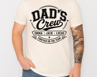 May include: A cream-colored short-sleeve t-shirt featuring a black graphic print. The design includes the text "DAD'S Crew" above the names "EMMA | JACK | LUCAS" and the phrase "FOREVER ON THE TEAM" with baseball-themed stitching accents.