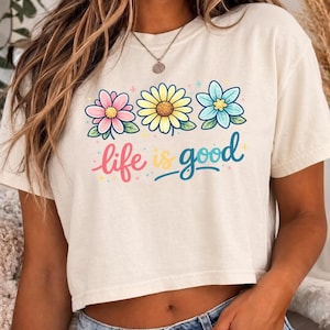 May include: Cream-colored crop top with a floral design and the phrase "life is good." The graphic features three flowers in pink, yellow, and blue, with green leaves. The text is in a colorful, playful font.