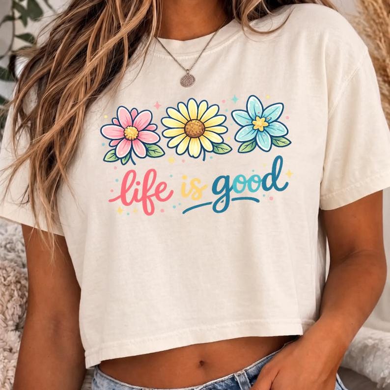 May include: Cream-colored crop top with a floral design and the phrase "life is good." The graphic features three flowers in pink, yellow, and blue, with green leaves. The text is in a colorful, playful font.