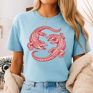 May include: Light blue t-shirt featuring a red illustration of two alligators in a circular design. The alligators are detailed with scales and open mouths. The shirt has short sleeves and a crew neck.