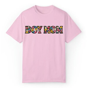 May include: A light pink t-shirt with the words "BOY MOM" in large, black outlined letters. The letters are filled with a colorful floral pattern, featuring pink, yellow, blue, and white flowers. The shirt has short sleeves and a crew neck.