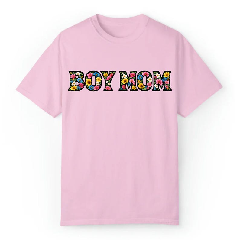 May include: A light pink t-shirt with the words "BOY MOM" in large, black outlined letters. The letters are filled with a colorful floral pattern, featuring pink, yellow, blue, and white flowers. The shirt has short sleeves and a crew neck.