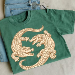 May include: Green crew neck t-shirt featuring a detailed illustration of two alligators facing each other. The alligators are in a light brown color. The shirt is paired with blue jeans and round eyeglasses.