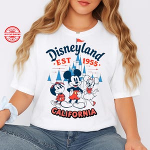 May include: White t-shirt with a Disneyland graphic. The design features the words "Disneyland EST 1955 CALIFORNIA" in navy blue, red, and white, alongside illustrations of Mickey Mouse, Minnie Mouse, and Pluto.