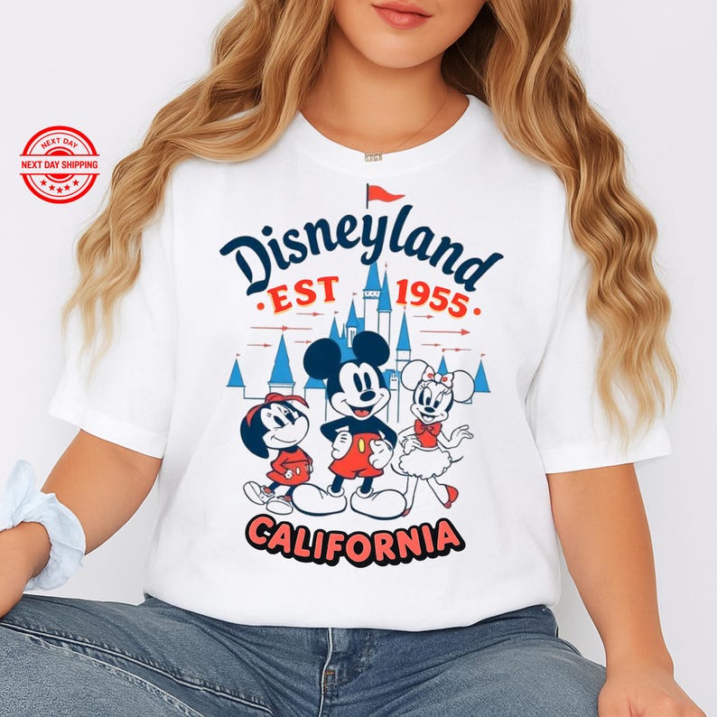 May include: White t-shirt with a Disneyland graphic. The design features the words "Disneyland EST 1955 CALIFORNIA" in navy blue, red, and white, alongside illustrations of Mickey Mouse, Minnie Mouse, and Pluto.