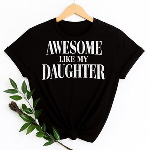 May include: Black t-shirt with the white text "AWESOME LIKE MY DAUGHTER." The shirt is hanging on a wooden hanger. A sprig of greenery is in the lower left corner.