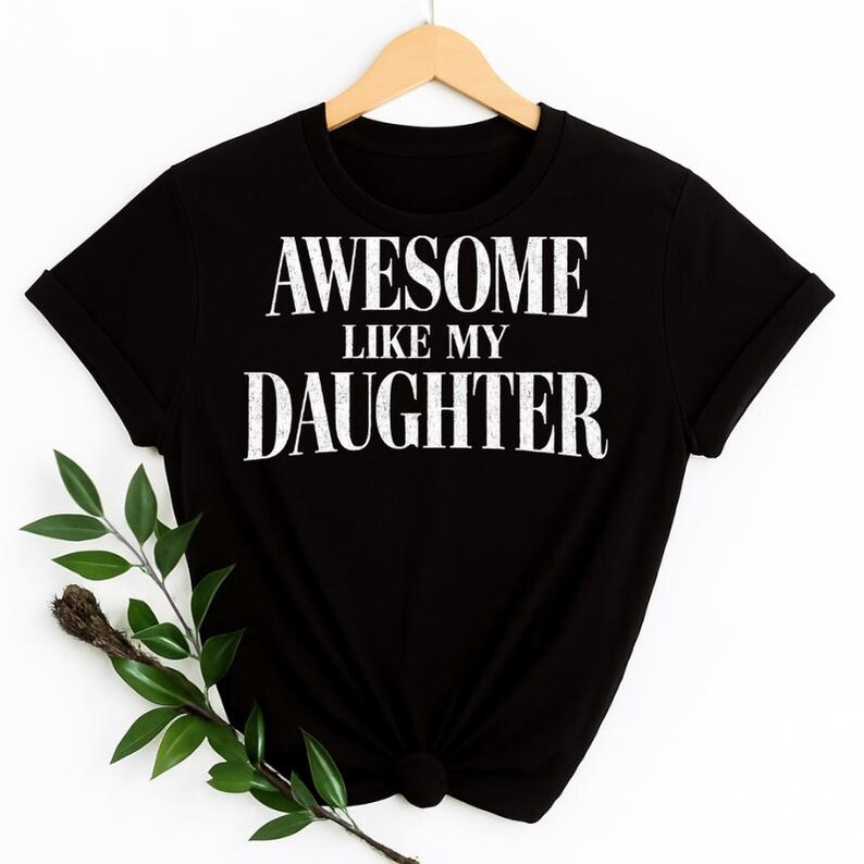 May include: Black t-shirt with the white text "AWESOME LIKE MY DAUGHTER." The shirt is hanging on a wooden hanger. A sprig of greenery is in the lower left corner.