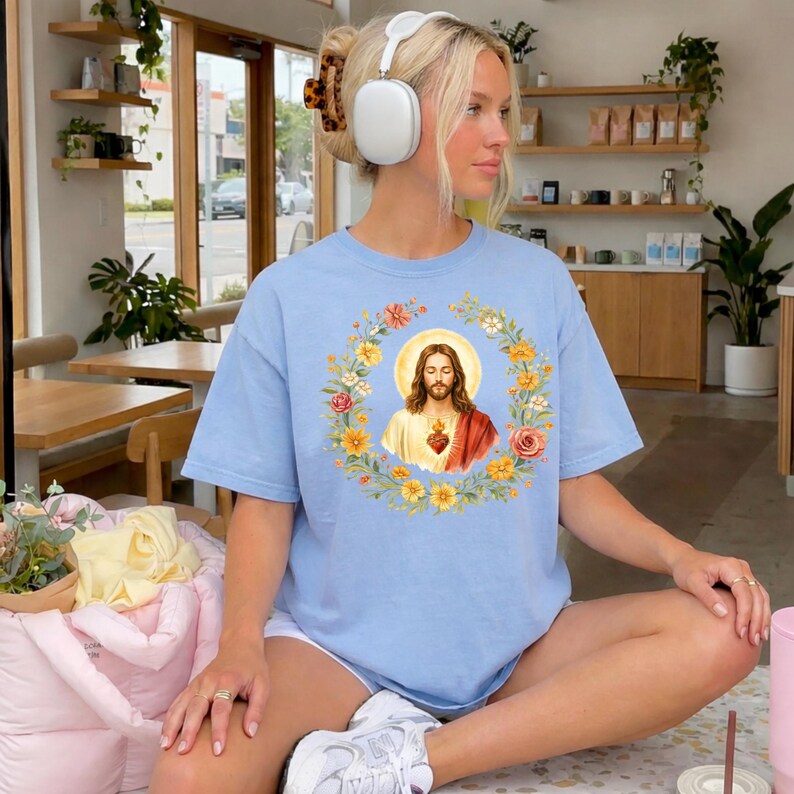 K&ouml;nnte beinhalten: Ein hellblaues T-Shirt mit einer zentralen Grafik von Jesus Christus mit heiligem Herzen, umgeben von einem farbenfrohen Blumenkranz aus gelben, orangen und rosa Bl&uuml;ten. Das T-Shirt hat kurze &Auml;rmel und einen Rundhalsausschnitt, getragen mit wei&szlig;en Kopfh&ouml;rern.