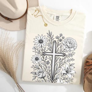 Op de afbeelding: Cr&egrave;mekleurig T-shirt met een zwart-wit bloemenkruisontwerp. Het shirt is omgeven door een strohoed, een gouden ketting en bruine sandalen. Het shirt heeft het merklabel "Comfort Colors".