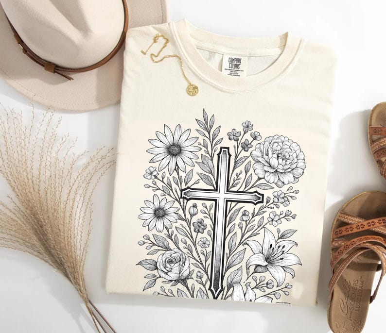 Op de afbeelding: Cr&egrave;mekleurig T-shirt met een zwart-wit bloemenkruisontwerp. Het shirt is omgeven door een strohoed, een gouden ketting en bruine sandalen. Het shirt heeft het merklabel "Comfort Colors".