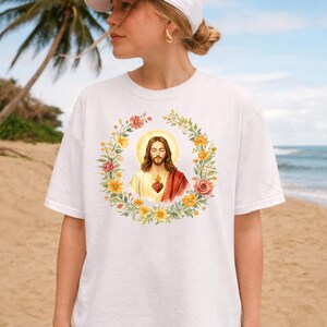 K&ouml;nnte beinhalten: Wei&szlig;es T-Shirt mit einer Aquarellillustration von Jesus Christus, umgeben von einem Blumenkranz. Das Bild zeigt Jesus mit Heiligenschein, bekleidet mit einem roten und wei&szlig;en Gewand und einem Herzsymbol. Der Kranz enth&auml;lt gelbe, rosa und orangefarbene Blumen.