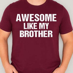 May include: A maroon t-shirt with the white text "AWESOME LIKE MY BROTHER" in a bold, sans-serif font. The shirt is a crew neck style and appears to be made of a soft material.