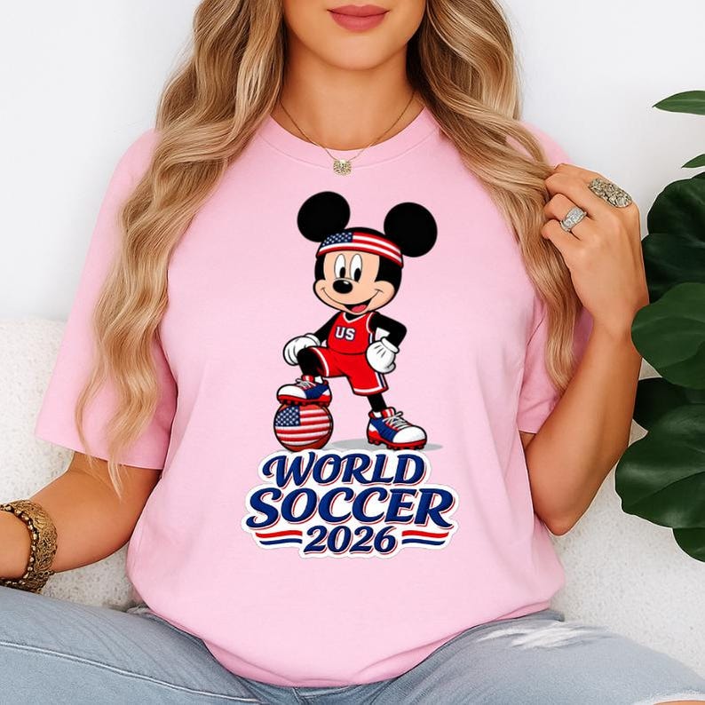 May include: Pink t-shirt featuring a graphic of Mickey Mouse in a soccer uniform with a soccer ball. The design includes the text "World Soccer 2026" in blue and red, with the US flag incorporated.
