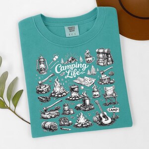 May include: Teal-colored t-shirt with a camping-themed graphic. The design features illustrations of camping gear, including a lantern, axes, a campfire, a guitar, and the words "Camping Life".