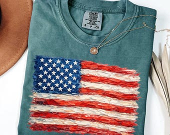 May include: A teal crewneck t-shirt featuring a textured, painted-style American flag graphic on the front. The shirt has a Comfort Colors brand label at the inner neckline, and a thin gold-tone chain necklace with a small circular pendant rests on top.