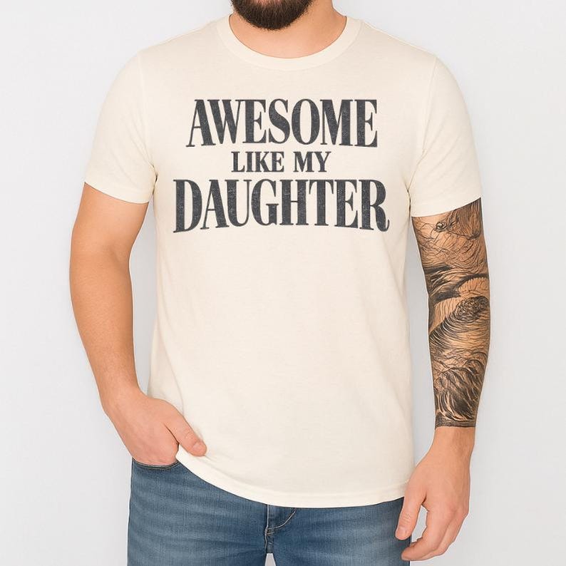 May include: A cream-coloured t-shirt featuring the text "AWESOME LIKE MY DAUGHTER" in a distressed black font. The short-sleeved shirt is worn with blue jeans.