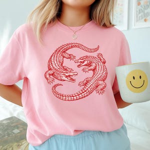 May include: A light pink t-shirt featuring a red illustration of two alligators facing each other. The alligators are detailed with scales and open mouths. The shirt is a crew neck style. A white mug with a yellow smiley face is held in the image.