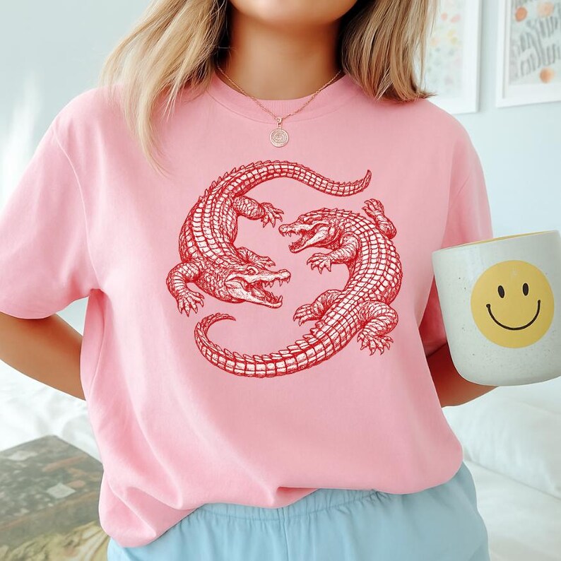 May include: A light pink t-shirt featuring a red illustration of two alligators facing each other. The alligators are detailed with scales and open mouths. The shirt is a crew neck style. A white mug with a yellow smiley face is held in the image.