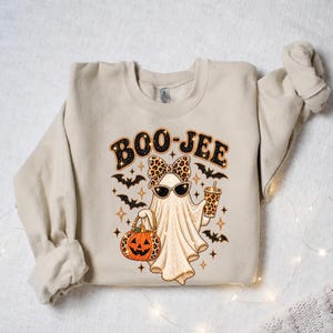 Pu&ograve; includere: Una felpa girocollo beige con la grafica di un fantasma che indossa un fiocco e occhiali da sole leopardati. Il fantasma tiene in mano una zucca e un bicchiere, circondato da pipistrelli e stelle. Il testo BOO-JEE &egrave; stampato sopra il fantasma.