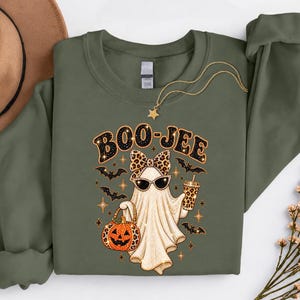 Pu&ograve; includere: Una felpa a maniche lunghe verde oliva con la grafica di un fantasma che indossa un fiocco e occhiali da sole leopardati, che tiene in mano una zucca leopardata e una bevanda. Il testo BOO-JEE &egrave; stampato sopra, circondato da pipistrelli.