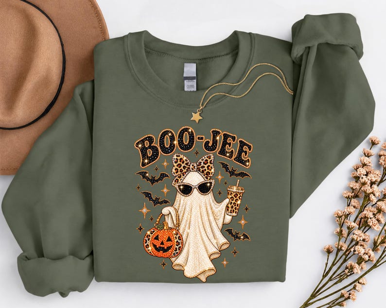 Pu&ograve; includere: Una felpa a maniche lunghe verde oliva con la grafica di un fantasma che indossa un fiocco e occhiali da sole leopardati, che tiene in mano una zucca leopardata e una bevanda. Il testo BOO-JEE &egrave; stampato sopra, circondato da pipistrelli.