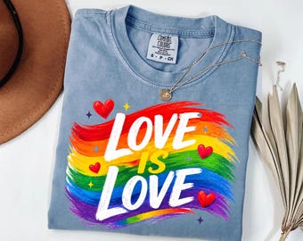 May include: A blue short-sleeve t-shirt featuring a graphic of a rainbow-colored brushstroke background with the white text "LOVE IS LOVE" and small red hearts and stars. The shirt has a Comfort Colors brand label at the inner neckline.