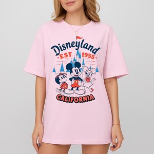 May include: A pale pink t-shirt with a Disneyland graphic. The design features Mickey Mouse, Minnie Mouse, and Goofy, with the words "Disneyland EST 1955 CALIFORNIA" in a whimsical font. The shirt has short sleeves and a round neck.