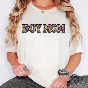 May include: Cream-colored t-shirt with the text "BOY MOM" in large, floral-filled letters. The flowers are a mix of pink, yellow, blue, and white. The shirt has short sleeves and a crew neck.