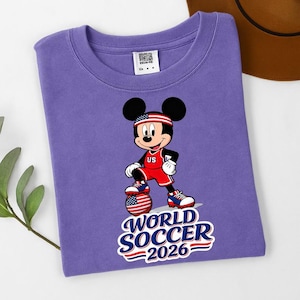 May include: A purple t-shirt featuring a graphic of Mickey Mouse in a soccer uniform with a soccer ball. The text "WORLD SOCCER 2026" is below the character. The character is wearing a red and white uniform with the letters "US".