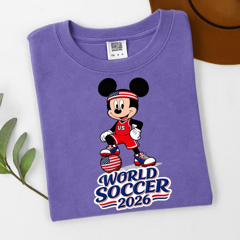 May include: A purple t-shirt featuring a graphic of Mickey Mouse in a soccer uniform with a soccer ball. The text "WORLD SOCCER 2026" is below the character. The character is wearing a red and white uniform with the letters "US".