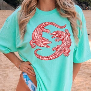May include: A light turquoise t-shirt featuring a red illustration of two alligators facing each other. The alligators are detailed with scales and open mouths. The shirt has short sleeves and a relaxed fit.