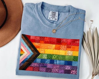 May include: A light blue crew neck t-shirt featuring a rectangular patch made of small, multi-colored fabric squares arranged in a pride flag pattern. The shirt has a Comfort Colors label at the neck and is displayed with a gold pendant necklace.