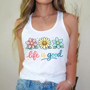 May include: White tank top with a floral design featuring three flowers in pink, yellow, and blue, with the words "life is good" in colorful script. The tank top is a casual, sleeveless style.