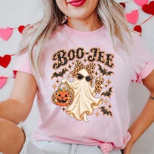 May include: A light pink t-shirt featuring a graphic of a ghost wearing sunglasses and a leopard print bow, holding a jack-o'-lantern and a drink cup. The text "BOO-JEE" appears above the ghost, surrounded by small black bats and star shapes.