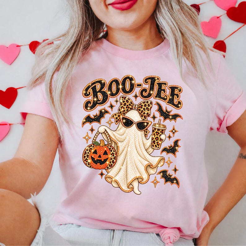 May include: A light pink t-shirt featuring a graphic of a ghost wearing sunglasses and a leopard print bow, holding a jack-o'-lantern and a drink cup. The text "BOO-JEE" appears above the ghost, surrounded by small black bats and star shapes.