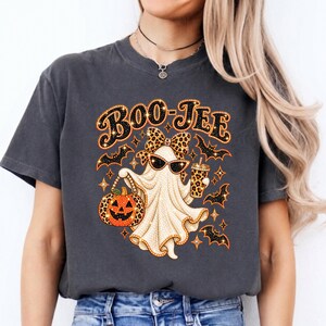 May include: A charcoal gray t-shirt featuring a graphic of a ghost wearing sunglasses and a leopard print bow. The ghost holds a leopard print pumpkin and a matching cup. The text BOO-JEE is printed above, surrounded by small bats and stars.