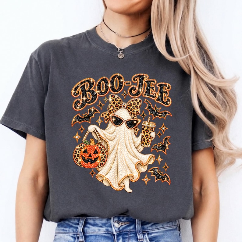 May include: A charcoal gray t-shirt featuring a graphic of a ghost wearing sunglasses and a leopard print bow. The ghost holds a leopard print pumpkin and a matching cup. The text BOO-JEE is printed above, surrounded by small bats and stars.