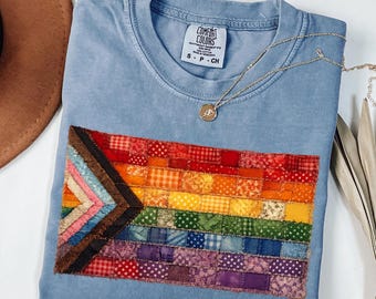 May include: A light blue crew neck t-shirt featuring a rectangular patch made of small, multi-colored fabric squares arranged in a pride flag pattern. The shirt has a Comfort Colors label at the neck and is paired with a thin gold chain necklace.