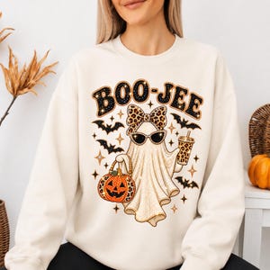 Pu&ograve; includere: Una felpa girocollo color crema con la grafica di un fantasma che indossa occhiali da sole e un fiocco leopardato. Il fantasma tiene in mano un secchiello a forma di zucca leopardato e un bicchiere. Il testo BOO-JEE &egrave; stampato sopra con pipistrelli.
