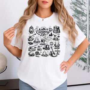May include: White t-shirt featuring a black and white camping-themed graphic. The design includes illustrations of a campfire, tent, guitar, and the text "Camping Life".