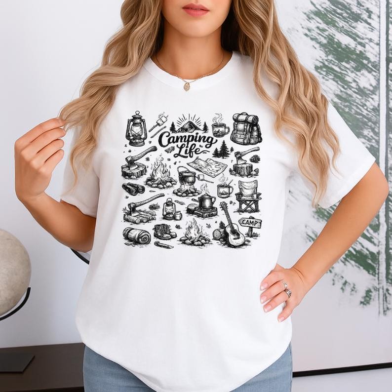 May include: White t-shirt featuring a black and white camping-themed graphic. The design includes illustrations of a campfire, tent, guitar, and the text "Camping Life".