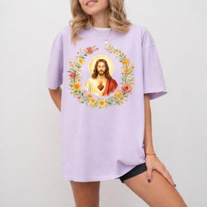 K&ouml;nnte beinhalten: Ein helllila T-Shirt mit einer Aquarellillustration von Jesus Christus, umgeben von einem Blumenkranz. Das Kunstwerk umfasst einen goldenen Heiligenschein und ein heiliges Herz. Das Shirt hat eine lockere Passform und kurze &Auml;rmel.