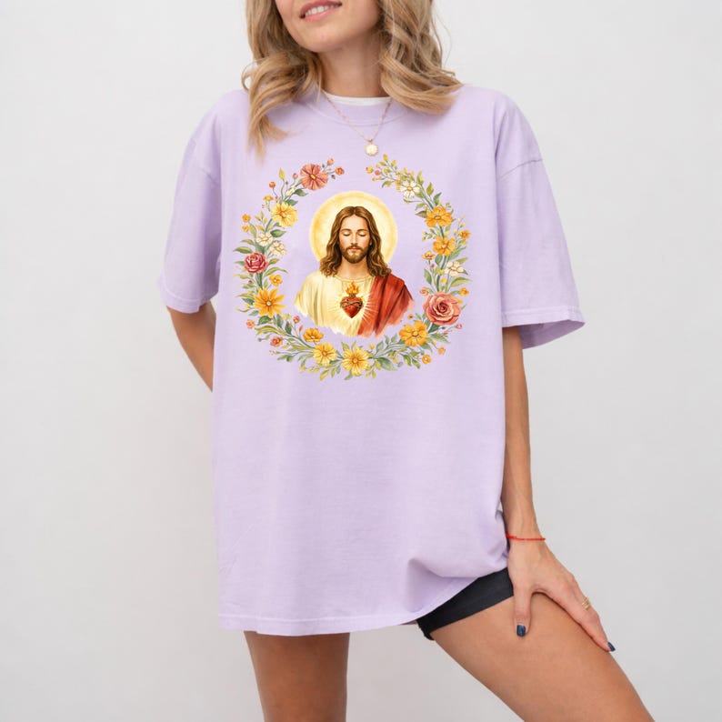 K&ouml;nnte beinhalten: Ein helllila T-Shirt mit einer Aquarellillustration von Jesus Christus, umgeben von einem Blumenkranz. Das Kunstwerk umfasst einen goldenen Heiligenschein und ein heiliges Herz. Das Shirt hat eine lockere Passform und kurze &Auml;rmel.