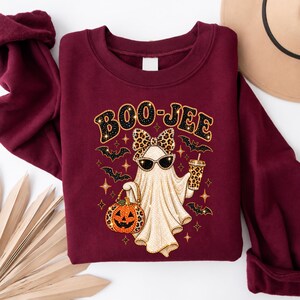 Pu&ograve; includere: Una felpa girocollo bordeaux con la grafica di un fantasma che indossa un fiocco e occhiali da sole leopardati. Il fantasma tiene in mano una zucca e un bicchiere abbinati. Il testo BOO-JEE &egrave; stampato sopra, circondato da pipistrelli e stelle.