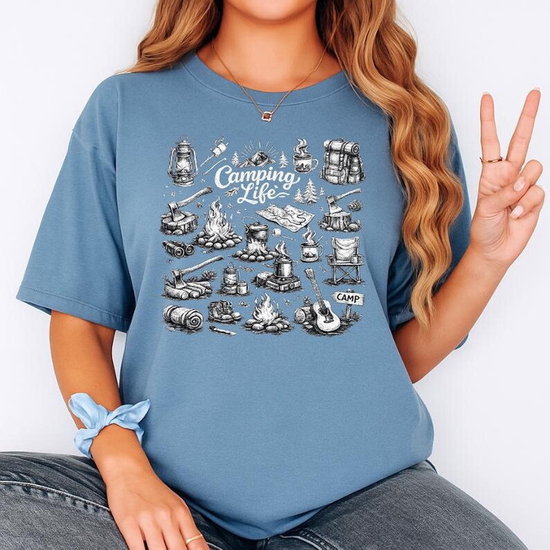 May include: A blue t-shirt with a camping-themed graphic. The graphic features camping elements like a campfire, lantern, guitar, and the text "Camping Life". The design is in black and white.