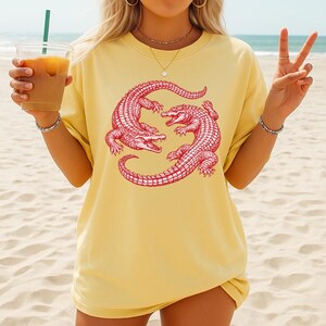 May include: Yellow t-shirt featuring a red graphic of two alligators in a circular design. The alligators are detailed with scales and open mouths. The shirt has short sleeves and a relaxed fit.
