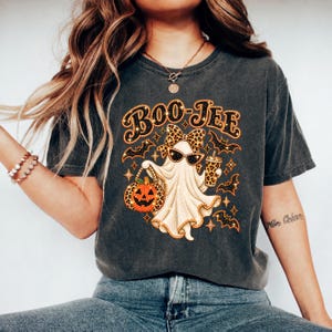 May include: A charcoal gray t-shirt featuring a graphic of a ghost wearing a leopard print bow and sunglasses. The ghost holds a pumpkin and a drink cup, surrounded by bats and stars. The text BOO-JEE is printed in a bold, glittery orange font.