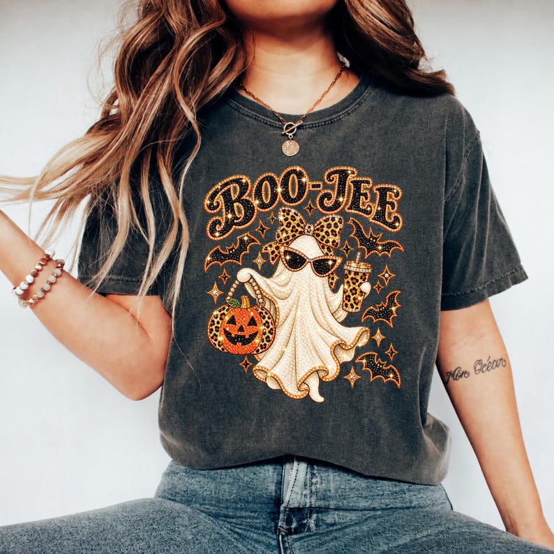 May include: A charcoal gray t-shirt featuring a graphic of a ghost wearing a leopard print bow and sunglasses. The ghost holds a pumpkin and a drink cup, surrounded by bats and stars. The text BOO-JEE is printed in a bold, glittery orange font.